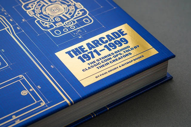 A close-up of a hardcover book titled "The Arcade 1971-1999" with a blue blueprint-style cover and a gold label, photographed on a dark gray surface.